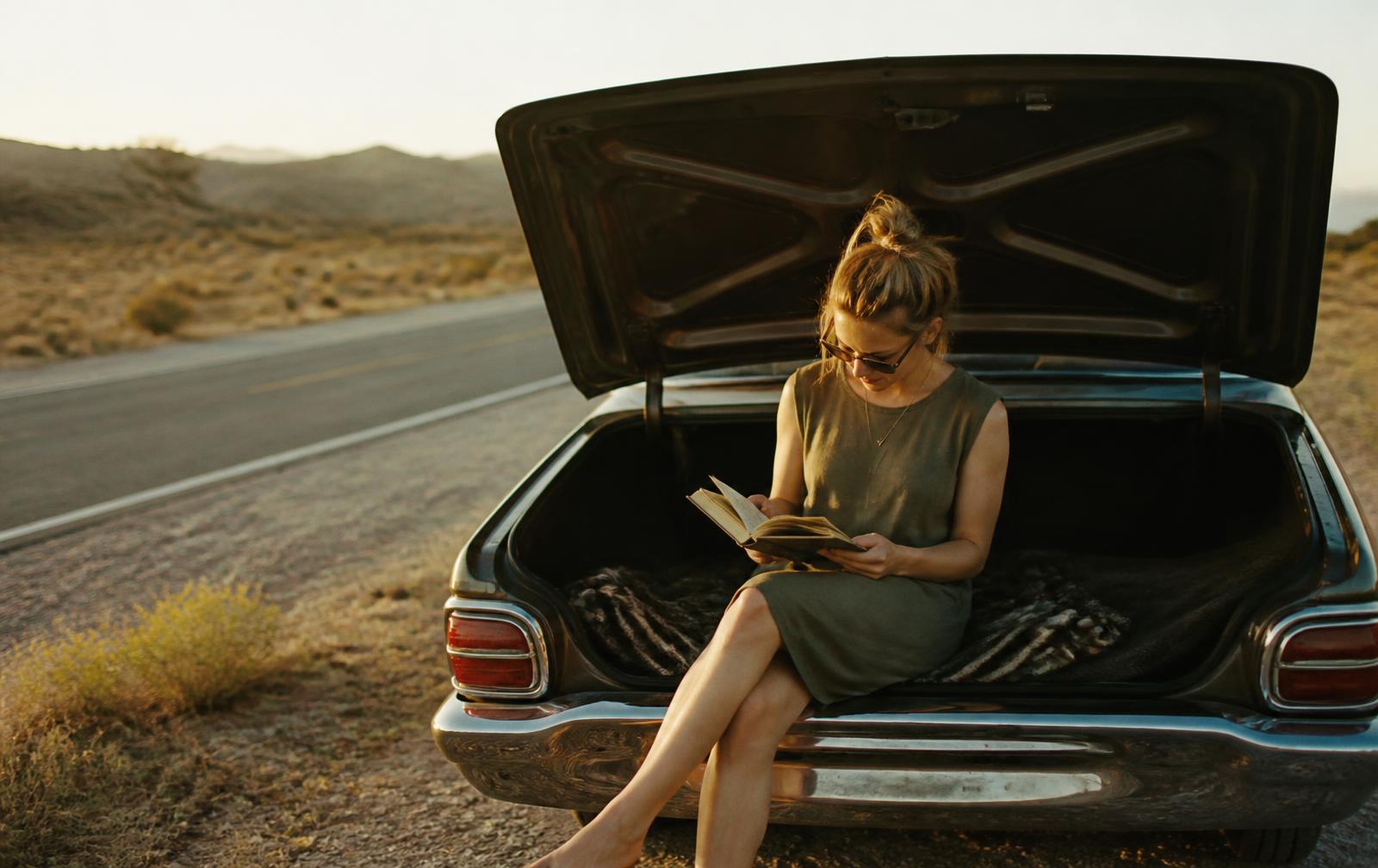 A woman reading a book in the open trunk of a car on a desert road at golden hour